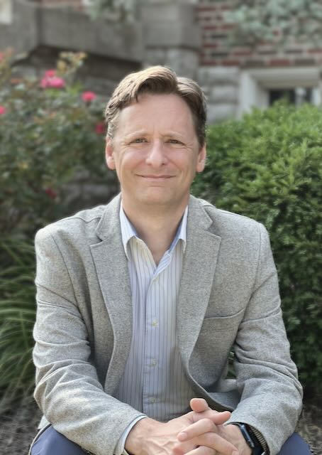Jason Sprang, mediator, seated with hands clasped in his lap wearing a gray blazer and striped shirt; garden courtyard with shrubs and a brick building in the background; calm, approachable, professional